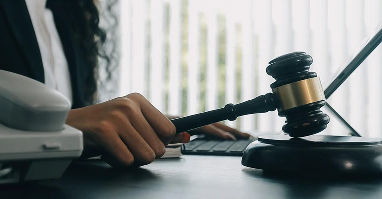 Lawyer’s hand holding a gavel next to a telephone and laptop on a desk in an office setting.