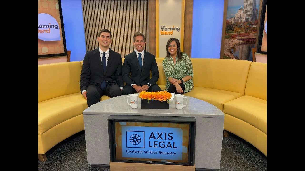 Three people—two men in dark suits and a woman in a green patterned blouse—are sitting on a yellow curved couch on the set of The Morning Blend. In front of them is a coffee table with two mugs, a flower arrangement, and a screen displaying the AXIS LEGAL logo and the tagline “Centered on Your Recovery.”