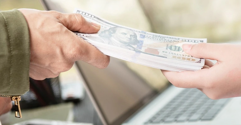 Close-up of a person handing over cash payment in front of a laptop.
