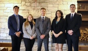 Five young professionals in business attire pose together in front of a cozy fireplace, exuding a warm and inviting atmosphere