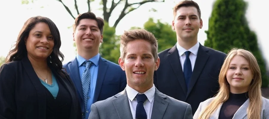 A group of five young individuals dressed in business attire, standing closely together in a professional setting.