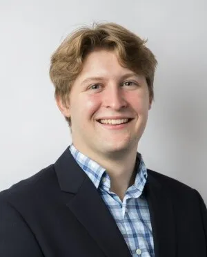 Portrait of a smiling young man with light brown hair wearing a dark blazer and blue checkered shirt.