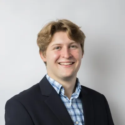 Portrait of a smiling young man with light brown hair wearing a navy blazer and blue checkered shirt.