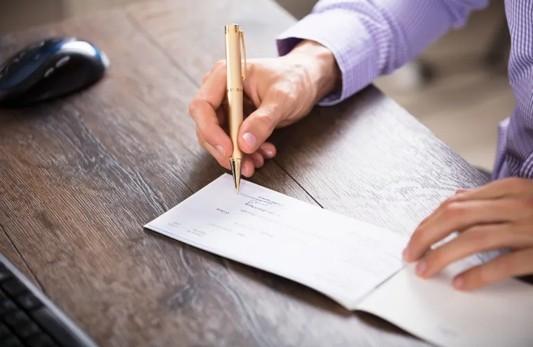 Person in a purple shirt signing a check with a gold pen on a wooden desk.