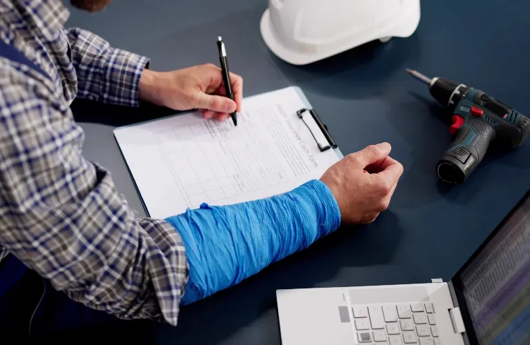 Man with blue arm cast filling out form on clipboard at desk with laptop, drill, and hard hat.