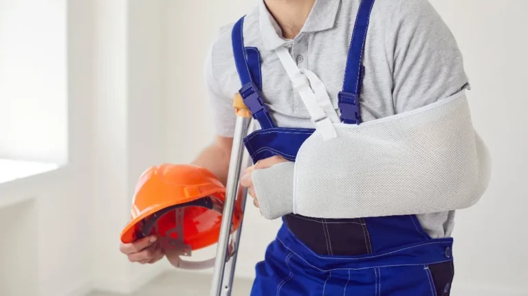 Man wearing blue overalls and gray shirt with arm in white sling, holding orange construction helmet and crutch.