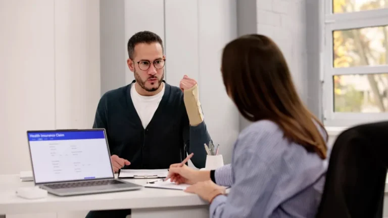 Two professionals discussing a health insurance claim, man with wrist brace explaining.