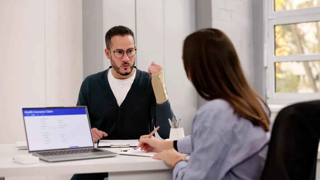 Two professionals discussing a health insurance claim, man with wrist brace explaining.